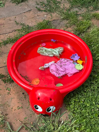 A red plastic sandbox, shaped like a ladybug, is filled with water and scattered with grass clippings and children's toys. Inside the sandbox are a pink shirt, a light green fabric item, a yellow watering can, a purple flower-shaped mold, and several small plastic cups and lids in orange and green. The sandbox is situated outdoors on a surface composed of both cracked concrete or paving stones and lush green grass. The surrounding area is also covered in grass and some scattered debris. The overall scene suggests a child's play area that has been left unattended, with elements of nature mixing with the man-made objects. The image appears to be taken from a high angle, looking down into the sandbox. The location is identified as Bolshie Ozery, Russia.