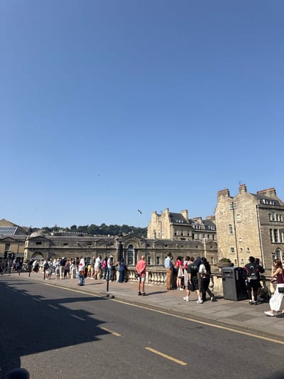The image depicts a sunlit outdoor urban scene on a clear day, likely in the city of Bath, United Kingdom. The vantage point appears to be from the eastern approach of Pulteney Bridge, looking west across the bridge itself.
In the foreground, a dark asphalt road occupies the lower left, marked with faded yellow double lines indicating a restricted area. Distinct shadows, presumably cast by objects or people outside the frame to the left, are visible on the road surface. Adjacent to the road on the right is a grey paved sidewalk.
A light-colored stone balustrade, characteristic of Pulteney Bridge, extends horizontally across the midground, separating the sidewalk from a lower area beyond. Approximately 25-35 individuals, appearing to be pedestrians or tourists, are congregated on the sidewalk and along the balustrade. Many are leaning on or looking over the balustrade, suggesting they are observing the view beyond the bridge. They are dressed in diverse casual summer attire. One man in a red shirt and light shorts stands facing left, with his back to the balustrade, near the center-right. A dark public rubbish bin is visible on the far right of the sidewalk, next to the balustrade. A slender dark pole stands vertically on the sidewalk near the center-left.
In the background, directly behind the balustrade, are several substantial stone buildings, characteristic of Bath's Georgian architecture, which are built directly onto the Pulteney Bridge. These buildings feature multiple windows, some with arched tops, and appear to house shops or residences. Beyond the bridge buildings, more traditional Bath stone structures with chimneys are visible, rising on a gentle incline. A line of dark green trees on a distant hillside forms the horizon line. On the far left, a structure with a glass roof is partially visible.
The sky is a uniform, clear light blue, occupying the upper two-thirds of the image. Several small, dark specks, likely birds, are visible in the sky. The scene is brightly illuminated by natural sunlight, casting discernible shadows and indicating daytime.