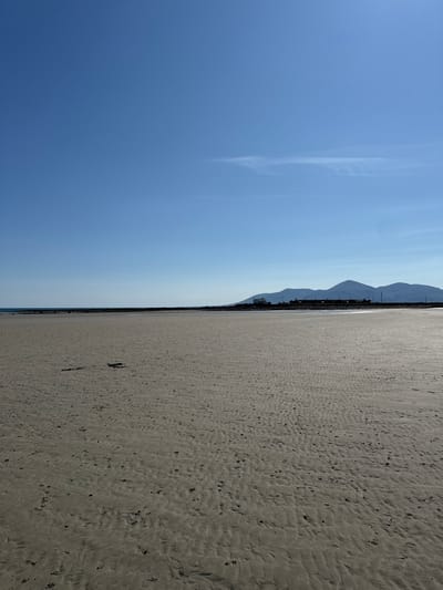The image depicts a wide, sandy beach on a clear, sunny day. The foreground is dominated by the textured sand, showing ripples from the receding tide and scattered dark debris. In the mid-ground, a low, dark line of rocks or a man-made barrier stretches horizontally across the frame, with the calm, blue sea visible beyond it. Further in the distance, a settlement of houses and buildings is silhouetted against a backdrop of rolling hills and mountains, suggesting a coastal town or village. The sky is a vast expanse of pale blue with a faint wispy cloud formation in the upper right quadrant. The location is identified as the beach in Ballykinler, United Kingdom. There are no people or animals visible in the scene.