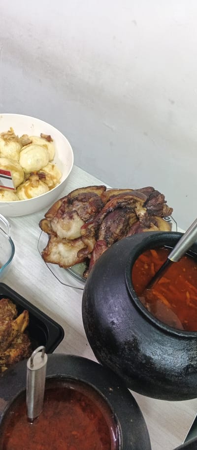 A close-up, overhead shot showcases a table laden with food, likely a home-cooked meal. In the foreground, two dark, rustic bowls with red stew and ladles rest on a light-colored wooden surface. To the right, a larger, black, earthenware pot filled with a reddish-orange soup, possibly borscht, is partially visible, with a metal ladle submerged within. To the left and center, a plate of succulent, roasted meat, appearing to be pork belly, is arranged. Behind the meat, a white bowl overflows with dumplings topped with fried onions and bacon bits. A small black container with a piece of fried meat is also visible on the table. The backdrop consists of a plain, light gray wall, suggesting an indoor setting. The scene is devoid of people, and the overall impression is one of abundance and hearty cuisine. This scene could be from the city of Ploske, Ukraine, given the traditional dishes.