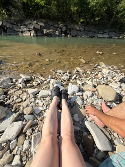 A low-angle, close-up shot shows the legs and feet of two individuals sitting on a rocky riverbank, with their legs extending towards the viewer. The person on the left wears black clogs and has bare legs. The person on the right has bare feet and legs, with one foot resting near the other individual's foot. The riverbank is covered in various sizes of light-colored stones. The river, with clear, shallow water revealing submerged stones and sand, flows behind them. The far bank of the river is lined with a broken, concrete embankment and dense green foliage. The scene appears to be in an outdoor, natural environment.