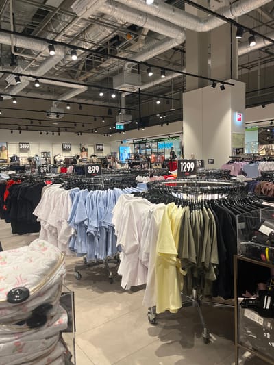 The image depicts the interior of a clothing store, possibly a fast-fashion retailer, with racks of t-shirts and other apparel prominently displayed. The store features a modern industrial design with exposed ductwork and track lighting on the ceiling. Several price tags are visible, indicating various prices such as 3499, 1899, 1999, 899, 1299, and 649, suggesting a range of price points for the merchandise. In the foreground, a quilted cosmetic bag with a floral pattern and gold zippers is displayed on a shelf. The store appears to be well-lit and organized, with a focus on presenting a wide selection of clothing. The location is specified as the city of Karavaevo, Russia. There are no people clearly visible in the image.