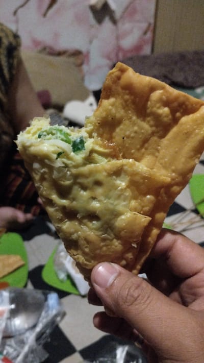 A person's hand holds a triangular pastry filled with scrambled eggs and green onions, with a bite taken out of it. The pastry is golden brown and appears to be fried. In the background, other pastries are visible on green plates, and a checkered blanket or tablecloth is laid out. A blurry figure of another person is in the far left background. The overall scene suggests a casual setting, possibly a meal or snack being shared. The lighting is dim, and the focus is primarily on the pastry. No visible text is present in the image. The location context provided is Sidodadi, Indonesia.