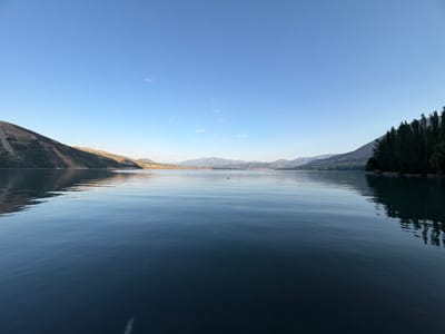The image shows a wide, calm body of water reflecting a clear blue sky. On the left, a barren, sun-drenched hillside slopes down to the water's edge, with several wind turbines visible on its upper reaches. The far side of the water is lined with distant mountains and a hazy, low-lying settlement. On the right, a densely forested hillside rises from the water, casting a shadow across the lake. The water itself is dark and smooth, with subtle ripples distorting the reflections of the surrounding landscape and sky. A small dark object, possibly a buoy, is visible in the distance on the water. The overall scene is one of natural tranquility and vastness, under a bright, cloudless sky.