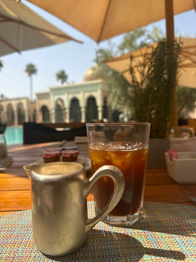 A glass of iced coffee sits on a colorful placemat on a wooden table. A silver creamer sits next to the glass. The coffee is filled with ice cubes. The setting is an outdoor patio with umbrellas providing shade. The patio is located at a resort in Ouled Jelal, Morocco, at coordinates 31.663, -7.872. The resort features a swimming pool in the background, and a large building with arched doorways can be seen. The building is painted in light tones, and the patio is situated in front of it. Lush green plants are also visible in the background. The scene is bathed in bright sunlight.