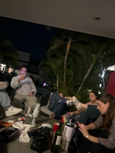 A group of people gather outdoors near palm trees at night. Some are sitting or lying down. There is a woman standing and interacting with someone below, with her foot near another person's face. There's a man sitting and holding what appears to be a cigarette. The area is littered with cups, bottles, and trash. A black bag is visible on the ground.