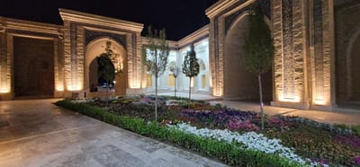 The image depicts the exterior of a building in Satkak, Uzbekistan, illuminated by artificial lighting at night. The architecture features large arched doorways and ornate tilework on the facades. In the foreground, a colorful flower bed is visible, planted with a variety of blooming flowers in shades of white, pink, purple, and yellow. Several trees, some with bare branches and others with dense foliage, are strategically placed within the courtyard. The ground is paved with large stone tiles. In the background, glimpses of the building's interior, including doorways, are visible through the arches. A few individuals can be seen in the distance, suggesting the area is accessible to the public. The overall impression is of a well-maintained and artistically designed outdoor space.