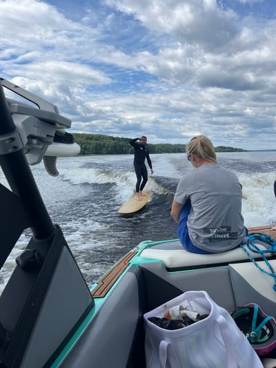 In Gromovo, Russia, on a body of water, a man in a black wetsuit is wakesurfing behind a boat. The man is standing on a yellow surfboard and is generating a significant wake. A woman in a grey t-shirt and blue pants is seated on the boat, facing away from the camera, observing the wakesurfer. The boat is partially visible in the foreground, and its wake is also evident. The background consists of a tree-lined shore and a cloudy sky. The image appears to be taken from another boat.