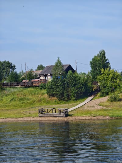 The image depicts a serene waterside scene in Galanino, Russia. In the foreground, a calm body of water reflects the sky and surrounding greenery. A small wooden dock juts out from the sandy bank into the water, equipped with simple seating. A paved path, possibly a walkway or ramp, leads from the bank upwards towards a slightly elevated area. The middle ground features a lush green grassy slope dotted with trees. A wooden boardwalk runs along the top of the slope, bordered by a railing. Behind the boardwalk, a cluster of houses with dark roofs and some exposed wooden structures is visible. Numerous trees, including birch and pine, are scattered throughout the landscape, some reaching considerable heights. The sky is a clear, pale blue with a few faint wispy clouds and a distant bird in flight. The overall impression is one of a peaceful, natural environment with some human infrastructure.