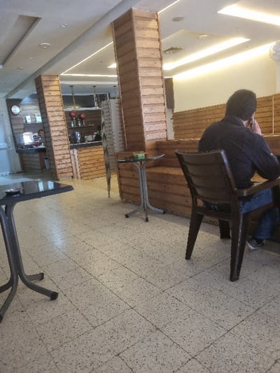 The scene takes place in a cafe with a tiled floor and wooden walls. A man sits on a wooden chair with a brown back and black seat, and he appears to be using a mobile phone. To the left of the man is a wooden banquette with a small, round wooden table. The man's legs are crossed, and he is wearing blue jeans and a dark shirt. The cafe has a small table in the foreground. The cafe is located in Tunis, Tunisia at the coordinates 36.838 latitude and 10.128 longitude. The lighting is bright. The cafe appears to be empty other than the man. The cafe has a ceiling with white tiles. The walls are made of wood. The walls are a light color, with a pale yellow hue. The walls are paneled. There is a clock on the wall, and a small counter to the left. The floor is a light gray, and covered in small black and white flecks, similar to terrazzo.