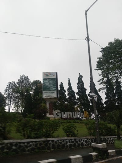 The image captures a roadside view of an agro-tourism destination on an overcast day. The scene is dominated by lush greenery, featuring various trees and bushes. In the center-left, a tall, vertical signboard stands on a stone base, providing information about the location. To the right of the signboard, large, silver-colored letters spelling "Gunung Ma" are arranged on a grassy slope, forming a prominent landmark.
The signboard, with a logo at the top reading "Agro Wisata N8," lists several amenities and services offered:
- RESORT
- CAMPING & GLAMPING
- BANQUET & P
- PUNCTION HALL (likely FUNCTION HALL)
- CATEHEST (likely CAFE & RESTO)
- OUTDOOR ACTIVITY
Below these, contact information is provided:
- OFFICE 24 HOURS
- 08112051616
- A partially visible website or email address starting with "www.agrowisat..."
Tall, slender trees, possibly conifers, line the landscape behind and to the right of the sign and sculptural text. Other trees with fuller, more rounded canopies are visible on the left and in the background. A dark, vertical utility pole with wires extends from the bottom right to the top center of the frame, partially obscuring some of the trees and the "Gunung Ma" text.
In the foreground, a dark asphalt road is visible, bordered by a low stone wall with a distinctive dark and light patterned texture. White and black striped barriers or curbs run along the edge of the road, suggesting a traffic lane. The sky is uniformly light gray and overcast, indicating either a cloudy day or low light conditions. The overall impression is one of a natural, serene environment, possibly an entrance to a park or resort, captured from a vehicle in motion, as suggested by slight motion blur on the road.