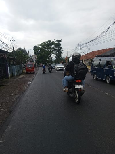 The image depicts a two-lane asphalt road, partially wet, under an overcast sky in Kamji Na-Yekha, Bhutan. In the foreground, a black automatic scooter carrying a driver and passenger is observed from behind. The driver wears a grey helmet, black hooded jacket, light blue jeans, and brown shoes, with a black backpack. The passenger wears light blue jeans and brown shoes. The scooter exhibits an illuminated red taillight and a license plate reading "AB 4701 RI." Ahead, two additional scooters, one dark-colored and one white, each with a helmeted rider, proceed down the road, followed by a white car. To the right, a blue minivan with a driver is in motion, paralleling the foreground scooter. The right side of the road features structures with reddish-brown tiled roofs and multiple utility poles supporting a dense network of overhead wires. On the left, a red minivan is parked on a dirt shoulder. Behind it, low concrete barriers, a dark metal fence, buildings with dark roofs, and a partial sign reading "ASIA TILE" are present. Utility poles with extensive overhead cabling and green foliage line the left roadside. Distant hills are faintly visible against the cloudy horizon.