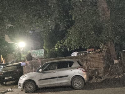 The image depicts a nighttime scene in Sector 12, Y-Block, Noida, India, specifically referencing house numbers 67-73 as indicated on a prominent green sign in the midground.
In the foreground, a silver Maruti Suzuki Swift hatchback is parked on the left side of a paved road, facing left. Its front left tire rests near some discarded white debris on the ground. Behind the Swift, a black SUV, likely a Maruti Suzuki Brezza, is partially visible, parked facing right. Its license plate, "UP16EA5751", is partially legible.
Above and behind the vehicles, a rough, weathered concrete or stone retaining wall spans horizontally across the midground. A large tree with dense green foliage rises from behind this wall, dominating the upper right and central portions of the image. Part of its thick trunk is visible on the right. Another white or light-colored vehicle is partially discernible behind the retaining wall, beneath the tree canopy.
A bright, yellowish-white street light illuminates the scene from the upper-left, with associated wires and poles visible. A smaller blue sign with Hindi text is partially obscured behind the black SUV. No individuals are present in the image. The scene is static, displaying parked vehicles and elements of an urban environment at night. The image exhibits noticeable digital noise, consistent with low-light photography.