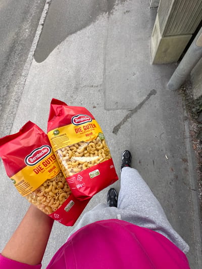 A first-person perspective image depicts an individual walking on a grey concrete or asphalt path in Grünwald, Germany. The person is wearing a bright pink top, grey sweatpants, and black athletic shoes with white soles and red accents. The individual's right arm and hand are visible, holding two identical red and yellow bags of "Bernbacher DIE GUTEN Hörnli" pasta, each containing 500g of elbow macaroni. The ground exhibits uneven coloration, with darker streaks and visible lines. To the right of the path, a light-colored rectangular concrete block structure is visible, alongside a grey downspout or pipe, with dark soil and sparse vegetation at the very edge.