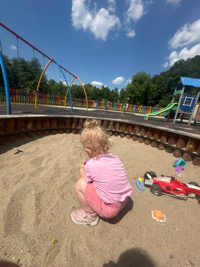 A young child with blonde hair in a bun, wearing a pink t-shirt and pink shorts, is playing in a sandbox in a park. The sandbox has a wooden border. In the sand, there is a red toy race car, a blue and yellow toy worm, a purple and teal toy, and an orange octopus toy. The park features swings, a slide, and a colorful fence. The scene is set on a sunny day with a blue sky and clouds.