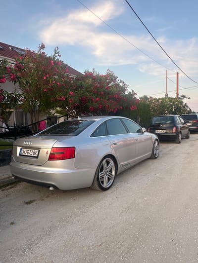An Audi A6 sedan, silver in color, is parked on a gravel road in front of a house. The car is facing right, with its rear toward the viewer. The license plate reads "CM 7073 BK". Pink oleander flowers are blooming on bushes in front of the house. Another black car and a dark SUV are parked behind the Audi. The sky is blue with some white clouds. The scene appears to be in a residential area, likely in the city of Chaidefto, Greece. There are no people visible in the image.