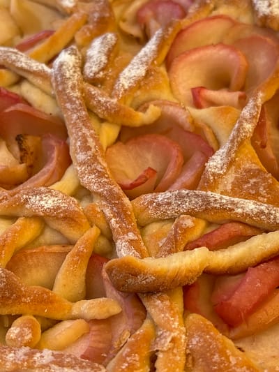 This is a close-up, overhead, vertical, eye-level shot of an apple tart. The tart has a lattice crust sprinkled with powdered sugar. The apples are sliced thinly and appear to be arranged in overlapping layers, with some showing a pinkish hue. The lighting is natural and bright, casting soft shadows and highlighting the textures of the crust and fruit. The scene is devoid of people or any environmental context, focusing solely on the baked good. The location in Вяйнянскі сельскі Савет, Belarus, cannot be determined from the image itself.