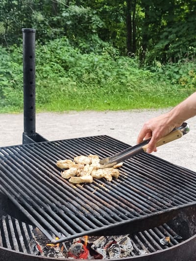 The image shows a close-up of a black metal barbecue grill in Slaka, Sweden. The grill is filled with charcoal embers that are glowing red. On the top grate of the grill, there are several pieces of raw chicken, appearing light in color. A person's hand, with visible veins and light skin, is holding a pair of metal tongs with wooden handles. The tongs are positioned over the chicken, with the metal tips gripping one of the pieces. The background consists of lush green foliage and trees, suggesting an outdoor setting. A gravel path is visible behind the grill. The overall scene depicts the preparation of food on a barbecue in a natural environment.