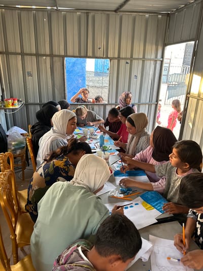 The image depicts an indoor art workshop held within a temporary structure composed of corrugated metal paneling, located in a-Zawayda, Palestinian Territory. Approximately 15 individuals, predominantly children and young women, are actively engaged in painting and drawing around two tables covered with white paper. Many participants wear hijabs.

Each individual possesses paintbrushes, small white cups containing water or paint, and sheets of paper displaying various artworks. Notable details include a painting with a horizontal blue stripe above a yellow stripe, suggestive of a flag. Pencils are also in use. A standing adult female wearing a patterned hijab observes the activity from the back-right.

The structure features a rectangular window opening on the left, looking out onto a blue chain-link fence; three children are visible observing the indoor scene through this opening. To the right, an open doorway reveals a bright, sunlit outdoor area with other structures and additional children observing or passing by. A metal shelf holding various containers is affixed to the left wall. Participants are seated on simple golden-colored chairs.
