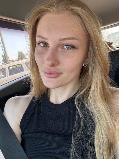 A young woman with blonde hair and green eyes is in the driver's seat of a car. She is looking at the camera and smiling. The car is parked on a street in Krasnyy Voskhod, Russia, during the day. The background shows trees and other cars. She is wearing a black tank top and a seatbelt.