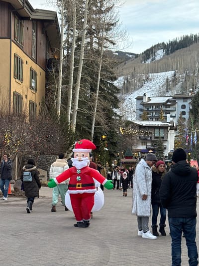 A vibrant scene displays an inflatable Santa Claus figure on a cobblestone street, surrounded by pedestrians in winter attire. The backdrop includes a building with windows, barren trees, and a snow-capped mountain range under a hazy sky. Strings of lights add a festive touch to the outdoor setting.