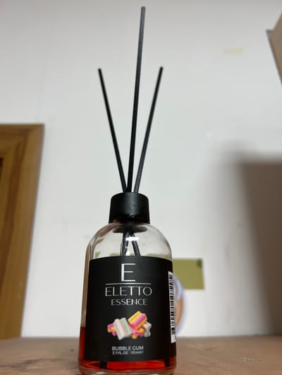 A close-up, eye-level shot shows a bottle of "Eletto Essence" reed diffuser with a "Bubble Gum" scent. The diffuser has a clear glass bottle filled with red liquid and a black cap with four black reeds inserted. The label on the bottle is black with white text and graphics, featuring the brand name "ELETTTO ESSENCE" and an image of colorful bubble gum pieces. The bottle rests on a wooden surface, and a light-colored wall is visible in the background. The image does not provide any information about the location in Kadamjay, Kyrgyzstan.