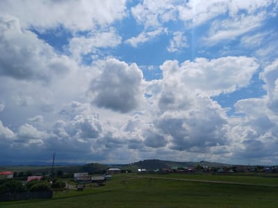 The image depicts a rural landscape under a partly cloudy sky, likely in or near Nigamatovo, Russia. In the foreground, a vast expanse of green grass stretches towards a cluster of buildings and trees. A dirt path runs through the grass, with tire tracks visible. Several structures, including houses with red roofs and what appear to be barns or outbuildings, are scattered across the mid-ground. A telecommunications tower stands near one of the buildings. The background features rolling green hills and a distant mountain range under a dramatic sky filled with large, white cumulus clouds interspersed with patches of blue sky. The overall scene suggests a serene, undeveloped rural setting. There are no people visible in the image.