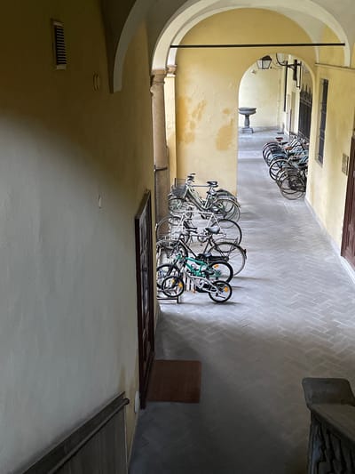 The image captures a view from a staircase looking down into a covered walkway in Piacenza, Italy. The walkway features a repeating pattern of arches, with a fountain visible in the distance through a further archway. The walls are painted a pale yellow, showing some signs of age and wear. Along the left side of the walkway, a series of parked bicycles are lined up against the wall. These range from adult-sized bikes to what appears to be a child's bicycle in the foreground. The floor is paved with a grey herringbone brick pattern. A dark wooden door is visible on the right side of the walkway, and a brown mat is placed on the floor in front of it. The lighting suggests it is daytime, with shadows cast by the arches and the bicycles. There are no people visible in the scene.