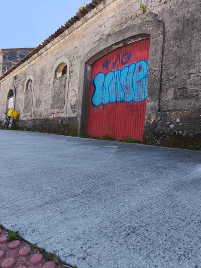 Captured in Cangas de Morrazo, Spain, the image presents a street scene. A gray stone building stretches along the left side, featuring a red-painted wooden door with blue graffiti and arched windows. A bicycle equipped with a yellow delivery bag is parked near the building. The foreground is dominated by a sloping gray concrete sidewalk with a red brick border visible at the bottom left. The sky above is light blue. The building's stonework shows signs of age and wear, with moss and discoloration present.