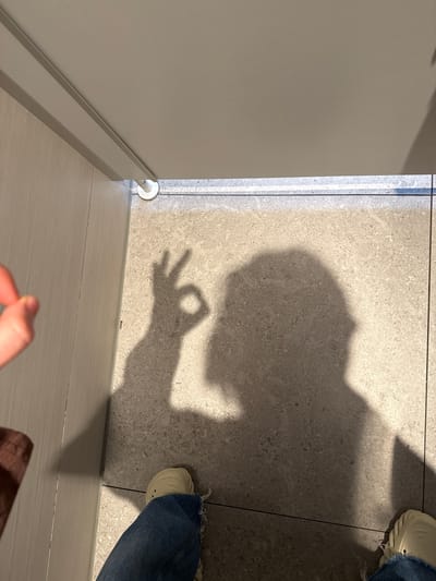 The image displays an indoor scene, likely in Waldshut, Germany, dominated by the shadow of a person cast upon a light-grey tiled floor. The shadow, occupying the upper right and central parts of the frame, distinctly shows the profile of a head and upper body, with one hand making an "OK" gesture.
To the right, the lower part of a person's leg clad in blue denim jeans and a light-colored, possibly beige, textured shoe is visible, partially obscured by the shadow. Another foot and leg are partially seen in the bottom right corner. The floor is covered with speckled, light-grey tiles with visible grout lines, suggesting a modern interior.
On the left side of the image, a plain light-grey wall stands vertically, meeting the floor with a matching baseboard. A small, white, circular object, possibly a floor-mounted doorstop or buffer, is visible where the wall meets the floor in the bottom left. In the immediate foreground, at the very bottom, out-of-focus fingers belonging to a hand are visible, suggesting the perspective of the photographer. A tiny metallic clip or staple lies on the lighter floor section near the baseboard.
The bright lighting creates sharp, well-defined shadows, indicating a strong light source, possibly natural light from a window or powerful artificial illumination. There is no visible text in the image. The scene captures a casual moment in what appears to be a contemporary building interior.