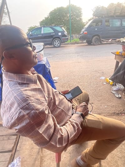 A man wearing glasses and a patterned shirt sits casually with his legs outstretched. He's holding a smartphone connected to a charger. Behind him is a street scene with several vehicles, including a van heavily loaded on its roof, parked along the roadside. Debris is scattered nearby, adding to the informal atmosphere.