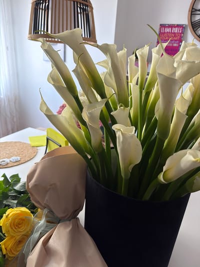 The image displays a vibrant bouquet of white Calla Lilies, occupying the majority of the frame. The lilies feature elegant, trumpet-shaped white blooms and prominent green stems. The bouquet is stylishly wrapped; the upper section is encased in black material, while the lower part is wrapped in a light brown, possibly fabric, material and tied with a light blue sheer ribbon.

Below the main bouquet, a smaller arrangement of fresh yellow roses is partially visible, providing a contrasting splash of color. The background consists of a plain, light-colored wall.

Several decorative objects are present in the background. On the left, a wooden object with horizontal slats, possibly a lampshade or basket, is visible. On the wall, a pink or purple sign is displayed, featuring text that reads: "LLAMA RULES," "LISTEN TO YOUR HEART," and "DO THE THINGS THAT MAKE YOU HAPPY." To the right, a circular woven mat with small white decorative elements is seen, along with a partial view of a bright yellow sticky note.

The scene suggests an indoor setting, likely a home or office, with natural, bright daytime lighting. The overall impression is one of floral beauty and personalized decor. The location context for this image is Cambrils, Spain.