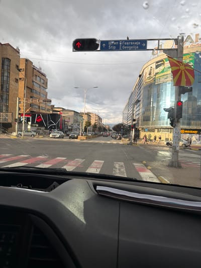 The image depicts a street scene in Strumica, North Macedonia, as viewed from inside a vehicle. A street sign indicates directions to Štip and Gevgelija, with the Macedonian flag displayed above it. A red traffic light with a left-pointing arrow is visible. The street has pedestrian crossings with red and white markings. Buildings line the street, including one with a Mozzart advertisement. Pedestrians are walking on the sidewalk. The sky is overcast.
