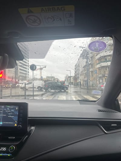 The image depicts an urban street scene on an overcast, rainy day, viewed from the interior of a vehicle. Raindrops are visible across the windshield.
**Vehicle Interior:**
The top portion shows the headliner with an 'AIRBAG' warning label. A purple 'CRIT'Air' environmental sticker is affixed to the upper right corner of the windshield. The vehicle's dashboard and infotainment system occupy the bottom third of the frame. The infotainment screen displays a navigation map, showing the vehicle's location near "Collège Jean Lurçat" and "Rue de la Prévoyance" in Vitry-sur-Seine, France. The climate control panel shows a temperature reading of "23.5".
**Exterior Scene - Foreground/Midground:**
The vehicle is stationary at a red traffic light, positioned directly behind a wet pedestrian crossing. A red traffic signal is visible to the left. A white panel van is stopped on the left side of the street, and a dark-colored sedan is visible in the center-midground, positioned within the intersection, potentially making a turn or stopped. The asphalt road surface is visibly wet from rain.
**Exterior Scene - Background:**
To the left, a modern, multi-story building with multiple windows is visible. In the central background, a large yellow tower crane indicates ongoing construction. To the right, a row of multi-story urban buildings with classical architectural elements line the street, receding into the distance. The sky is uniformly gray and overcast. No individuals are clearly identifiable within the scene.