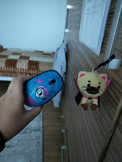 A hand holds a blue and pink computer mouse with a cartoon face design, standing in front of a wood-grain cabinet with a small yellow cat plush toy hanging from the top. The cat is wearing a pink cape and has black eyes and a black nose. There is a bed with a checkered brown blanket behind the hand and a white sweater hanging from the cabinet. The location is Florești, Romania.
