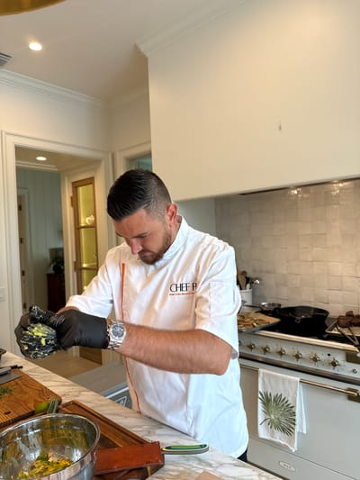In a brightly lit, upscale kitchen in East Naples, Florida, a male chef, identified by the "CHEF P" logo on his white chef's coat, is actively preparing food. He wears black gloves and a silver wristwatch, and is seen vigorously mashing avocado in his gloved hands, which are held over a stainless steel mixing bowl containing more avocado. The kitchen features a marble countertop, wooden cutting boards, and a large white gas range with brass knobs. A decorative kitchen towel with a palm frond design hangs from the oven door. The background reveals a doorway leading to another room with wooden cabinets and a glimpse of a dining area. The overall scene suggests a professional culinary environment within a residential setting.