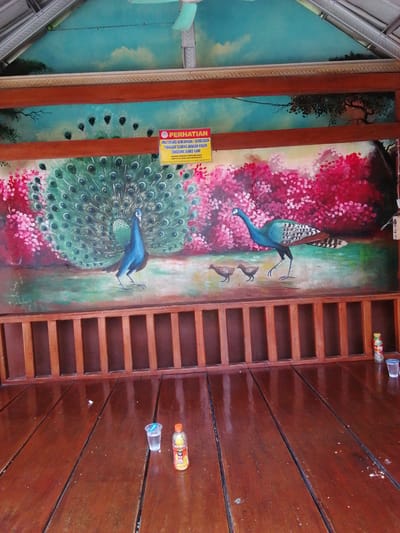 The image shows a mural of peacocks and chicks on a wall, located in a covered outdoor seating area in Tegalwangi, Indonesia. The mural features a large peacock with its tail feathers spread, a smaller peacock to the right, and three chicks below it. The background of the mural depicts a sky with clouds and trees. In front of the mural is a wooden bench with vertical slats. On the wooden floor in front of the bench, there are two small plastic cups, one with liquid and one empty, and a bottle of beverage. A yellow sign with red text is attached to the mural, stating "PERHATIAN" (Attention) and providing rules or notices, partially obscured by the wooden structure. The overall scene suggests a public or semi-public space, possibly a restaurant or rest area, decorated with art.