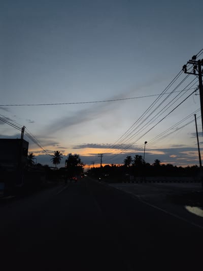 The image depicts a road scene at dusk in Keudealuerheung, Indonesia. The road stretches forward in perspective, flanked by vegetation and buildings in silhouette. Power lines crisscross the sky above the road. The sky exhibits a gradient of colors from gray and blue at the top to orange and yellow at the horizon, indicating a setting sun. The road surface is mostly dark, obscuring details. Distant vehicles are visible on the road with their headlights on. A puddle is visible on the right side of the road.