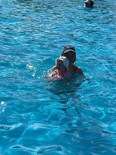 In the city of Koloksha, Russia, a young woman wearing a blue baseball cap and a black swimsuit is in a swimming pool, holding a toddler. The toddler is wearing a white cap and a pink swimsuit. Both are partially submerged in the clear blue water, with ripples and sunlight reflecting on the surface. Another person, wearing a sun visor, is visible swimming in the background of the pool. The scene depicts a recreational activity in a public swimming facility.
