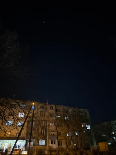 A nighttime exterior scene in Ciorescu, Moldova, captures a multi-story residential apartment building beneath a clear, dark sky. The sky exhibits numerous visible stars, including a prominent, bright celestial object (likely a planet) in the upper-left quadrant and the recognizable constellation Orion in the upper-right. A tall utility pole with a single illuminated yellow streetlamp stands in the left-center foreground, partially obscured by the bare, leafless branches of a tree extending from the left. The apartment building, approximately five to six stories high, features a light-colored facade with numerous windows, many of which are illuminated with artificial light. The ground floor appears to house a brightly lit commercial establishment, possibly a shop, with several individuals congregated outside and multiple parked vehicles nearby. A second, similar apartment building is partially visible in the far right background. No specific discernible actions or interactions are evident beyond general presence and lighting.