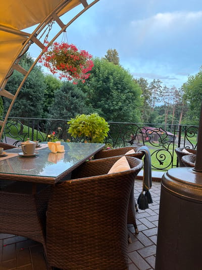 The image depicts an outdoor seating area at a restaurant or cafe, likely located in Izmailovo, Russia, given the presence of a dacha-style building in the background and lush greenery characteristic of the region. The scene is set on a patio paved with rectangular stone tiles. In the foreground, a glass-topped table is set with a teapot, cups, and a napkin holder, suggesting a tea or coffee service. Wicker chairs with cushions surround the table. Above the seating area, a fabric awning is suspended by a metal frame, and a hanging basket filled with red flowers dangles from it. Beyond the patio, a decorative wrought-iron railing overlooks a grassy area with trees and a rustic wooden building with a red roof. The sky is overcast, with a soft, diffused light. No people are visible in the frame.