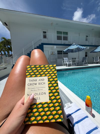 This image captures a first-person perspective of an individual relaxing by a swimming pool on a clear, sunny day in Lauderdale-by-the-Sea, United States.
In the foreground, the lower torso and upper thighs of a person with tanned skin are visible, wearing a black swimsuit bottom. The individual's left hand, with light pink polished fingernails, holds an open copy of the book "Think and Grow Rich" by Napoleon Hill, which features a cover design of repeating yellow gold bars on a dark green background. A white towel with two prominent blue horizontal stripes is draped over the person's right leg. Adjacent to the towel, on the white poolside coping, rests a red and orange bottle with a bright yellow cap.
The midground is dominated by a clear, blue swimming pool with sunlight reflecting off its rippled surface. Beyond the pool, a two-story building, painted white with distinct blue trim and railings, stands under a vivid blue sky dotted with scattered white clouds. The building features white staircases leading to the second-story balcony, which is lined with white railings. Directly adjacent to the pool, several outdoor seating arrangements consisting of grey chairs with white frames are positioned around white tables, shaded by two large blue and white striped umbrellas. To the far left of the building, a white cabinet-like structure with multiple compartments is discernible.
In the background, dense green foliage, including prominent palm trees, is visible on the left side, indicating a tropical or subtropical setting. The scene is well-lit by natural sunlight, casting visible shadows.