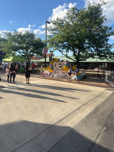 In Des Plaines, Illinois, on a sunny day with scattered clouds, a lively street scene unfolds. A large, illuminated sign spells out "DESPLAINES," adorned with a cascade of colorful balloons in shades of gold, purple, and blue. Several people are present, including individuals taking photos of the sign. A woman in a black dress is captured mid-stride, holding a camera. To the left, a group of three women and two children are also taking pictures. Further back, green tents suggest a marketplace or event setup. An American flag is visible on a flagpole, and trees provide dappled shade. The ground is paved with concrete and brick, and metal barriers line the right side of the image.