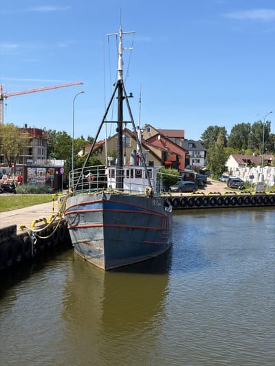 The image depicts a sunny day in Łeba, Poland, likely along its waterfront. In the foreground, a weathered, dark blue fishing boat is docked, its bow facing the viewer. The boat has a red trim along its gunwale and a nameplate that reads "DR Słava". Yellow ropes secure the boat to the dock, which is lined with tires.
Behind the boat, a walkway runs alongside the harbor. A small building with colorful advertisements is visible on the left, and further back, buildings with red-tiled roofs line the street. A large orange construction crane dominates the background on the left, indicating ongoing development. On the right side of the harbor, a line of large, conical concrete structures stands, possibly breakwaters or mooring elements. Trees and a clear blue sky with wispy clouds complete the scene. Several cars are parked along the street behind the buildings. There are no people clearly visible or engaged in any activity in this specific shot.
