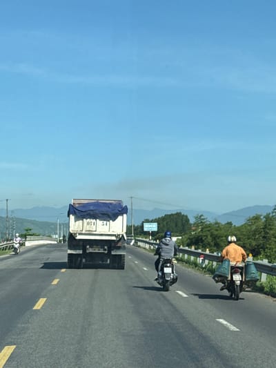 On a clear, sunny day, a truck with a blue tarp covers its load drives on a paved road. The truck is white with the license plate 074 51. Two men on motorcycles follow behind the truck. The first motorcycle is ridden by a person wearing a gray jacket and a blue helmet. The second motorcycle is ridden by a person wearing an orange shirt and a white helmet, with two large baskets attached to the rear of the motorcycle. The road is lined with a metal railing and trees on the right side. In the distance, mountains are visible under a clear blue sky. The scene appears to be on National Route 1A in Diên Lạc, Vietnam.