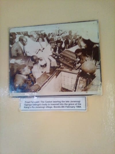 An aged, sepia-toned photograph depicts a solemn outdoor funeral service, mounted on a light-colored wall. In the foreground, a dark, rectangular casket is being lowered into a rectangular grave opening by several individuals. Numerous adult males, predominantly dressed in suits or formal attire, densely surround the grave, observing the interment process. Some individuals are directly involved in handling the ropes or straps used to descend the casket, while others stand as spectators. The background shows additional gathered people, contributing to a crowded scene.
A caption below the photograph identifies the event: "Final Farewell: The Casket bearing the late Jaramogi Oginga Odinga's body is lowered into the grave at his Kang'o Ka Jaramogi village, Bondo. 5th February 1994." Kang'o Ka Jaramogi village in Bondo is located within the broader region of South Sakwa ward, Kenya.