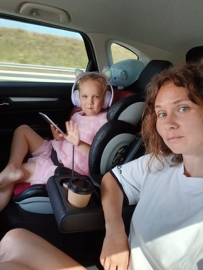 The image shows two females in the back of a car, presumably traveling. On the left, a young girl with blonde hair is seated in a car seat and wearing headphones. She is holding a tablet in her left hand and waving with her right. She is wearing a pink dress. To her right, an adult female with curly brown hair is visible, looking towards the camera. She is wearing a white t-shirt. In the foreground, a cup holder with a disposable cup is visible. Outside the car window, a blurry landscape of dry grass and a highway can be seen. The scene is lit by natural sunlight.