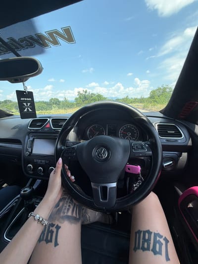 An eye-level, slightly tilted shot captures the interior of a car, likely a Volkswagen due to the steering wheel logo, set against a bright, sunny outdoor scene in Belovitsa, Bulgaria. The image is taken from the driver's perspective, focusing on the steering wheel, dashboard, and the driver's hands and legs.
The driver's right hand, adorned with a silver charm bracelet and manicured nails with pink polish, grips the bottom of the steering wheel. A prominent black tattoo reading "1980" in an old English font is visible on the driver's right thigh, while another tattoo is partially visible on the left leg. A bright pink car key is attached to the steering column, hanging near the indicator stalk.
The dashboard features a speedometer at zero, suggesting the car is stationary, and an infotainment system below the central air vents. A black air freshener, branded "areon -VERSION- X", hangs from the rearview mirror. Through the windshield, a clear blue sky with scattered white clouds is visible, along with green trees and foliage in the distance, indicating a rural or scenic setting. Reflections of text, possibly "NM", are seen on the upper left of the windshield.
Visible text includes "1980" (tattoo), "areon -VERSION- X" (air freshener), "PASSENGER AIRBAG OFF" (dashboard warning light), "OK" (button on steering wheel), and "AIRBAG" (on the steering wheel). The overall impression is a casual moment, perhaps paused before or during a drive on a clear day.