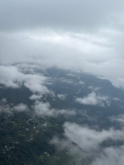 The image depicts a mountainside scene in Khumbupasanglahmu, Nepal, partially obscured by heavy fog and clouds. The terrain is rugged and forested, with a small village or cluster of buildings visible amidst the trees. The mountain's peak is shrouded in mist. The overall atmosphere is hazy and overcast.