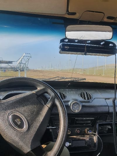 The scene is captured from the interior of a vehicle, presenting a view of an outdoor environment in Egizbulak, Uzbekistan. Inside the vehicle, a black four-spoke steering wheel, textured and bearing a "GRAND PRIAM" emblem, dominates the left foreground. The black, textured dashboard is visible, featuring air vents, a circular analog clock with a black face and silver bezel, an audio system, and a 12V charger emitting a blue light. A black rectangular device is suspended directly below the rearview mirror by black cords. The overhead lining and beige sun visors are also visible. Outside, a dirt road extends into the distance under a clear, light blue sky. To the right of the road, a green wire mesh fence runs parallel, behind which numerous light-colored utility poles or support structures are arranged. On the left side, beyond the fence line, several large, light-colored skeletal structures, possibly solar panel mounts or construction frames, are present within a flat, arid landscape. The lower portion of a person's leg, clad in khaki-colored trousers, is partially visible below the steering wheel.